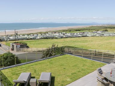 A garden with two sun loungers and a dining table overlooking a coastal area with caravans and a beach at Melin Y Plas in Llanaber