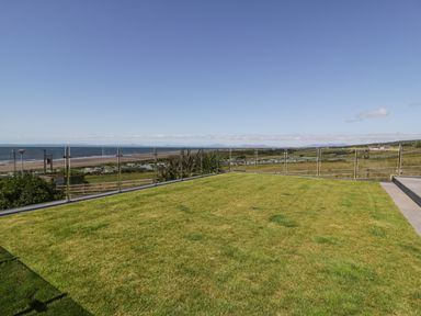 A grassy patio area with glass fencing overlooking a beach and distant hills at Melin Y Plas in Llanaber