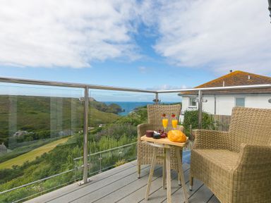 A balcony with wicker chairs and a small wooden table with drinks and food overlooking green hills and the ocean at Rowena in Mullion