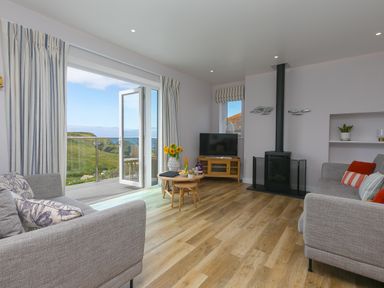 A living room with two grey sofas, a round wooden coffee table, a television on a wooden stand, a wood-burning stove, and open doors leading to a balcony with a view of green hills and the sea at Rowena in Mullion