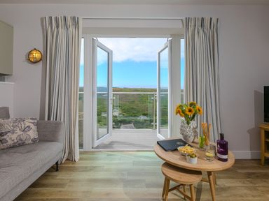 A living room with a grey sofa and open double doors leading to a balcony overlooking a green landscape at Rowena in Mullion