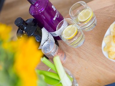 Two glasses with lemon slices on a wooden table with bottles of gin and tonic water and a bowl of potato chips