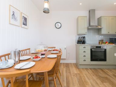 A dining area with a wooden table set with plates cups strawberries and cake beside a kitchen with green cabinets and stainless steel oven at Rowena Mullion