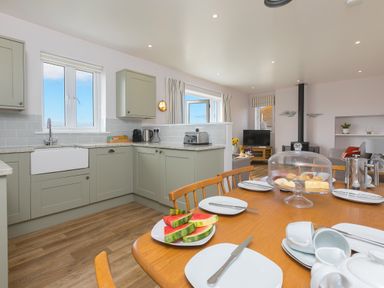 A kitchen and dining area with a wooden dining table set with plates cups and knives and watermelon slices on a plate at Rowena in Mullion