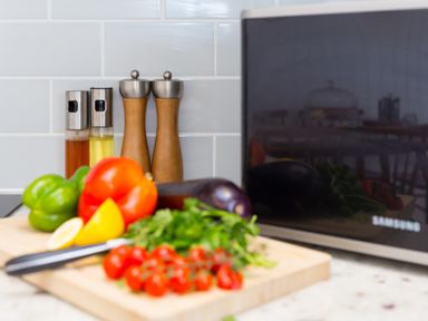 A kitchen countertop with a cutting board holding various vegetables and herbs next to a microwave and oil and pepper shakers at Rowena in Mullion