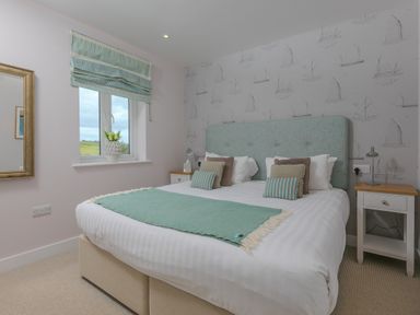 A bedroom with a large bed with pillows and a green throw by a window with green curtains and bedside tables at Rowena in Mullion