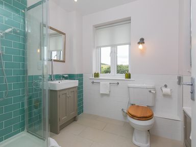 A bathroom with a shower, a sink with cabinet, a toilet with wooden seat, a window and a wall light at Rowena in Mullion