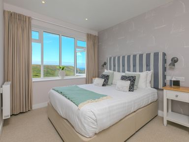 A bedroom with a large bed with striped headboard cushions a window with curtains and a view of green hills at Rowena in Mullion