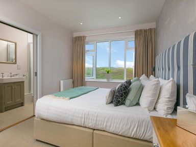 A bedroom with a bed with striped headboard and pillows near a window with curtains and a view of hills at Rowena in Mullion