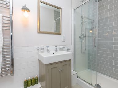 A bathroom with a sink and cabinet a mirror a light fixture a glass door shower and bottles of toiletries at Rowena in Mullion
