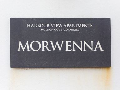 A black sign with white text reading Harbour View Apartments Mullion Cove Cornwall Morwenna