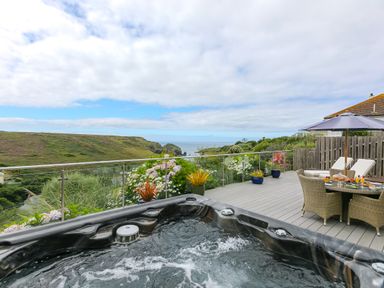 An outdoor deck with a hot tub a dining table with chairs and an umbrella overlooking hills and the sea at Morwenna in Mullion