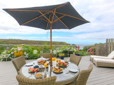 An outdoor patio with a round table set with food and drinks under a large umbrella overlooking greenery and water at Morwenna in Mullion