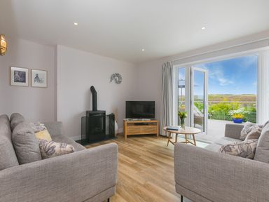 A living room with two sofas a coffee table a TV and a stove next to open doors leading to a balcony at Morwenna in Mullion