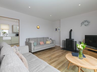 A living room with two grey sofas a wooden coffee table with flowers a black stove and a television at Morwenna in Mullion
