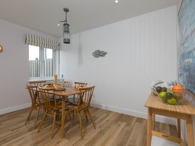 A dining room with a wooden table and six chairs with food and drinks on the table a bowl of fruit on a side table and a window with striped curtains at Morwenna in Mullion