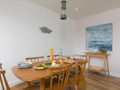 A dining room with a wooden table and chairs a table with food and glasses and a side table with fruit and a painting at Morwenna in Mullion
