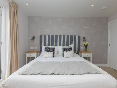 A bedroom with a bed with striped headboard two bedside tables and floral wallpaper at Morwenna in Mullion