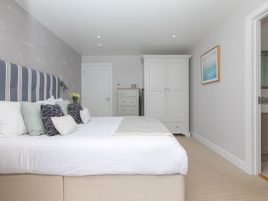 A bedroom with a bed with multiple pillows a chest of drawers a wardrobe and a door leading to a bathroom at Morwenna in Mullion
