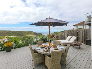 An outdoor deck with a round table and chairs under an umbrella with food and drinks on the table and two lounge chairs against a wooden fence at Morwenna in Mullion