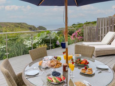 A patio dining table set with breakfast items and an umbrella with lounge chairs and garden view at Morwenna in Mullion