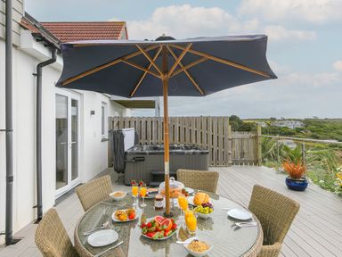 An outdoor patio with a glass table set with breakfast food and drinks beneath a large umbrella with wicker chairs and a hot tub behind the table at Morwenna in Mullion