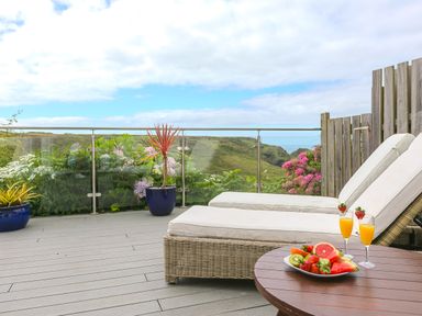 An outdoor deck with two cushioned lounge chairs a round wooden table with fruit and drinks and plants along a glass railing at Morwenna in Mullion