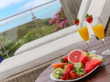 A wooden table with a plate of sliced fruit and two glasses of orange juice with strawberries on a patio at Morwenna in Mullion