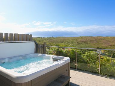 A bubbling hot tub on a deck overlooking green hills with a glass railing at Verity in Mullion