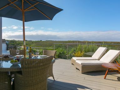 A patio with wicker chairs and a table under an umbrella and two lounge chairs overlooking greenery at Verity in Mullion