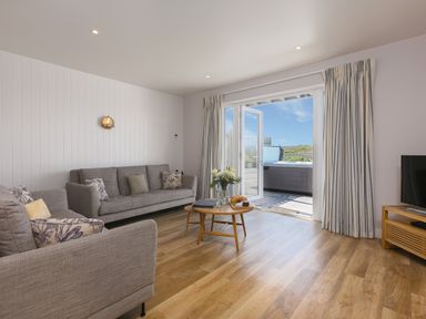 A living room with two gray sofas a round wooden coffee table with flowers and snacks and open doors leading outside at Verity in Mullion
