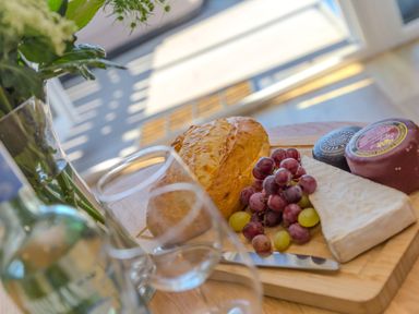 A wooden board with a loaf of bread grapes three types of cheese and a knife on a table with two empty glasses and a bottle near a window at Verity in Mullion