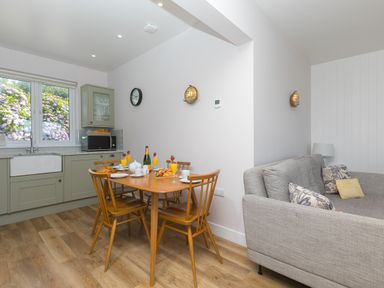 A kitchen and dining area with wooden chairs a table set with breakfast items a grey sofa and a window overlooking flowers at Verity in Mullion