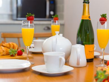 A kitchen table set with glasses of orange juice garnished with strawberries a teapot a milk jug a bottle of champagne croissants and plates at Verity in Mullion