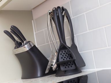 A kitchen counter with a black knife block and black kitchen utensils on a stand against a tiled wall at Verity in Mullion