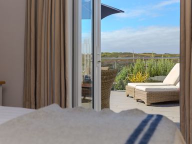 A bedroom with beige curtains and a bed looking out to a patio with wicker chairs and a sun lounger at Verity in Mullion