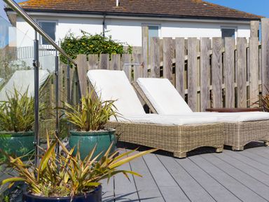 Two wicker lounge chairs with white cushions on a wooden deck next to potted plants and a glass railing in an outdoor patio area at Verity Mullion