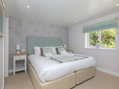 A bedroom with a double bed, bedside tables with lamps, patterned wallpaper, a window with a roman blind and a vase with flowers at Verity in Mullion