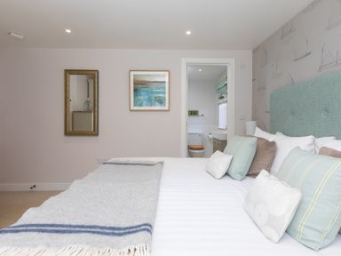 A bedroom with a bed, cushions, a wall mirror, a framed picture and an open door to a bathroom at Verity in Mullion