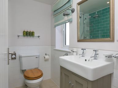 A bathroom with a toilet with a wooden seat a sink with two taps a mirror and a window with a blind at Verity in Mullion