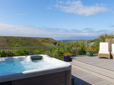 An outdoor deck with a hot tub, wicker chairs, potted plants, and a glass railing overlooking green hills and the sea at Verity Mullion
