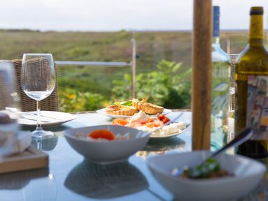 An outdoor dining table with plates of food wine bottles and an empty wine glass with a view of greenery at Verity in Mullion
