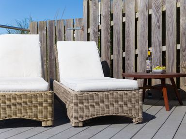 Two wicker sun loungers with white cushions beside a wooden table with a bottle of wine glasses and a bowl of grapes on a deck at Verity in Mullion