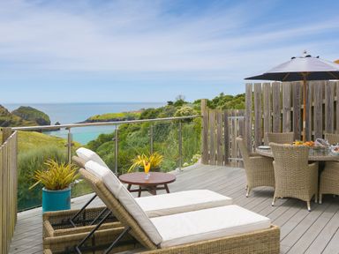 An outdoor deck with wicker sun loungers and a dining table under an umbrella overlooking coastal cliffs and the sea at Daphne in Mullion
