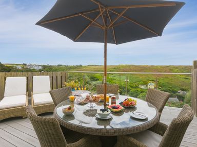 A round glass outdoor dining table with wicker chairs and a large umbrella on a deck with breakfast items and a view of green hills and houses at Daphne in Mullion