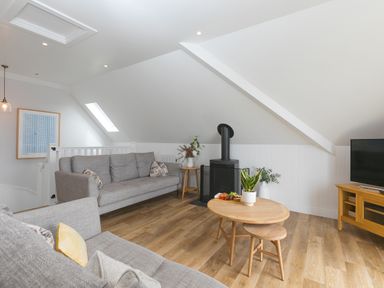 A living room with two grey sofas a wood-burning stove a round wooden coffee table with a plant a TV on a wooden stand and a framed picture on the wall at Daphne in Mullion