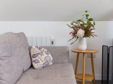 A grey sofa with a patterned cushion next to a wooden side table holding a vase of flowers at Daphne in Mullion