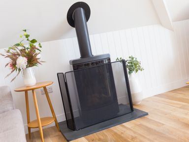 A wood burning stove with a safety screen on a wooden floor with a wooden side table and flowers at Daphne in Mullion
