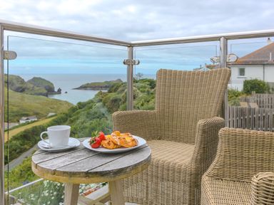 A wooden table with a cup and a plate of pastries and strawberries on a balcony with wicker chairs and a sea view at Daphne in Mullion