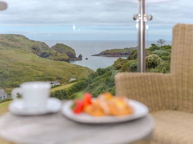 A coastal view with hills and a body of water seen through glass near a table with a cup and plate and a wicker chair at Daphne in Mullion